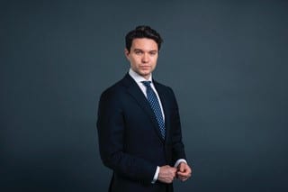 Sam wears a suit on a dark grey background