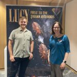 Two people standing next to a banner which reads: Lucy, first to the Trojan asteroids