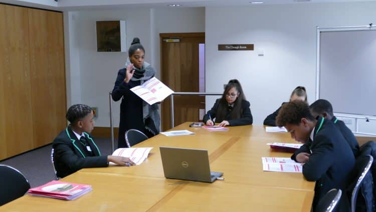 Postgraduate student giving a tutorial lesson to five year 9 pupils, who are seated around a rectangular table, looking at booklets.