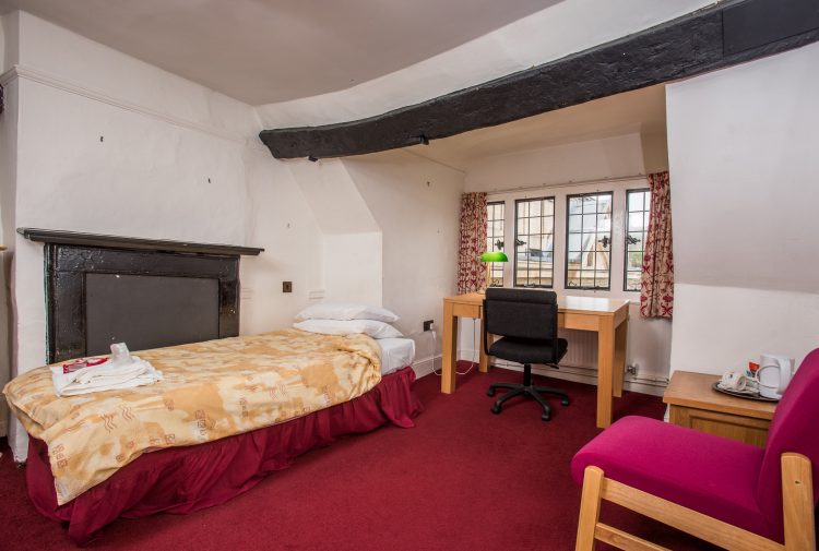 Staircase 4, Room 5. A red carpet and curtains around a mullioned window. Above the window is a historic wooden beam. The bed is next to a historic fireplace, and a red chair and desk beneath the window are also pictured.