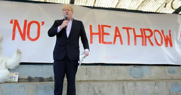 Boris Johnson with a microphone in hand stands on a stage in front of a sign saying No Heathrow