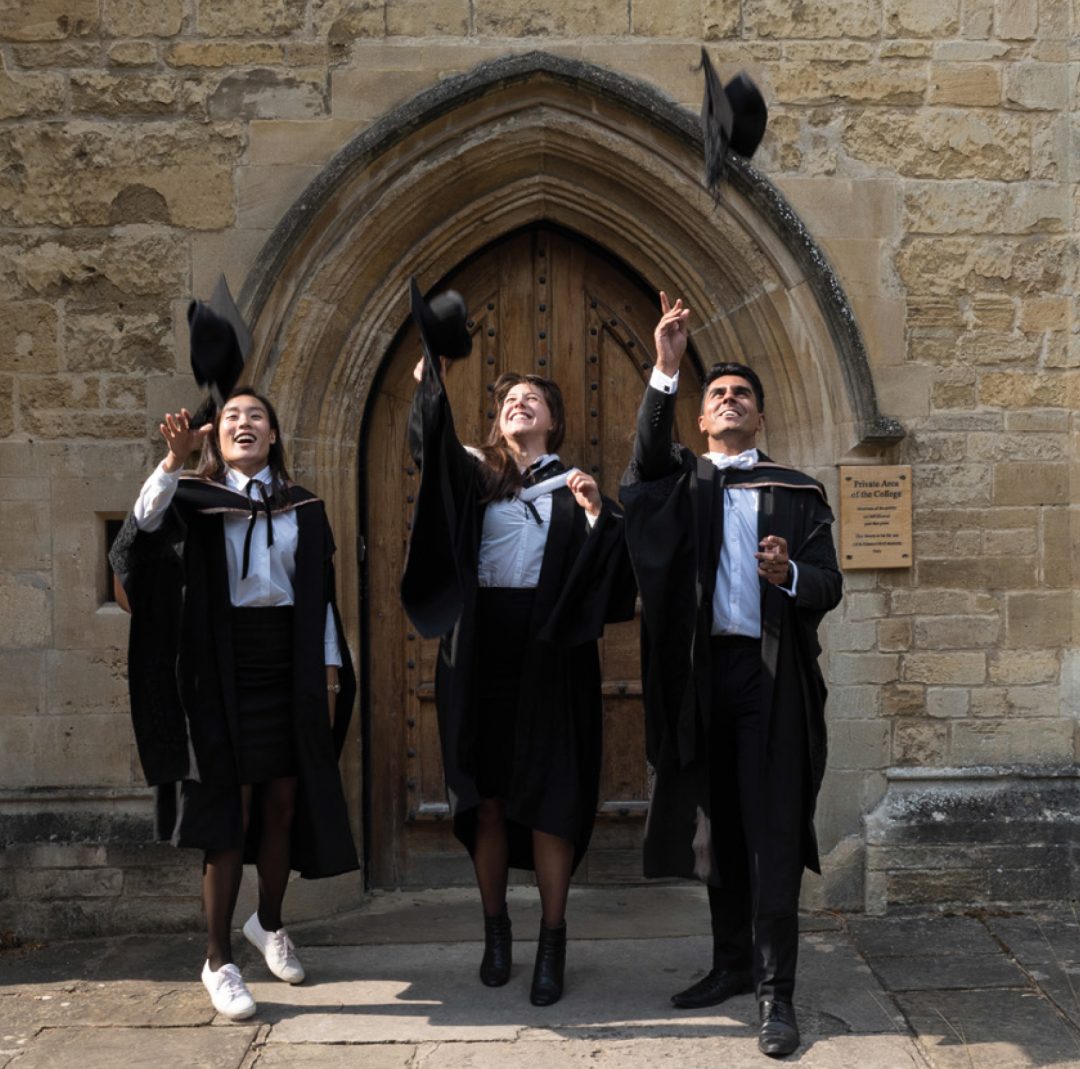 Graduates throwing their mortar boards in the air in front of the doors to the Teddy Hall library